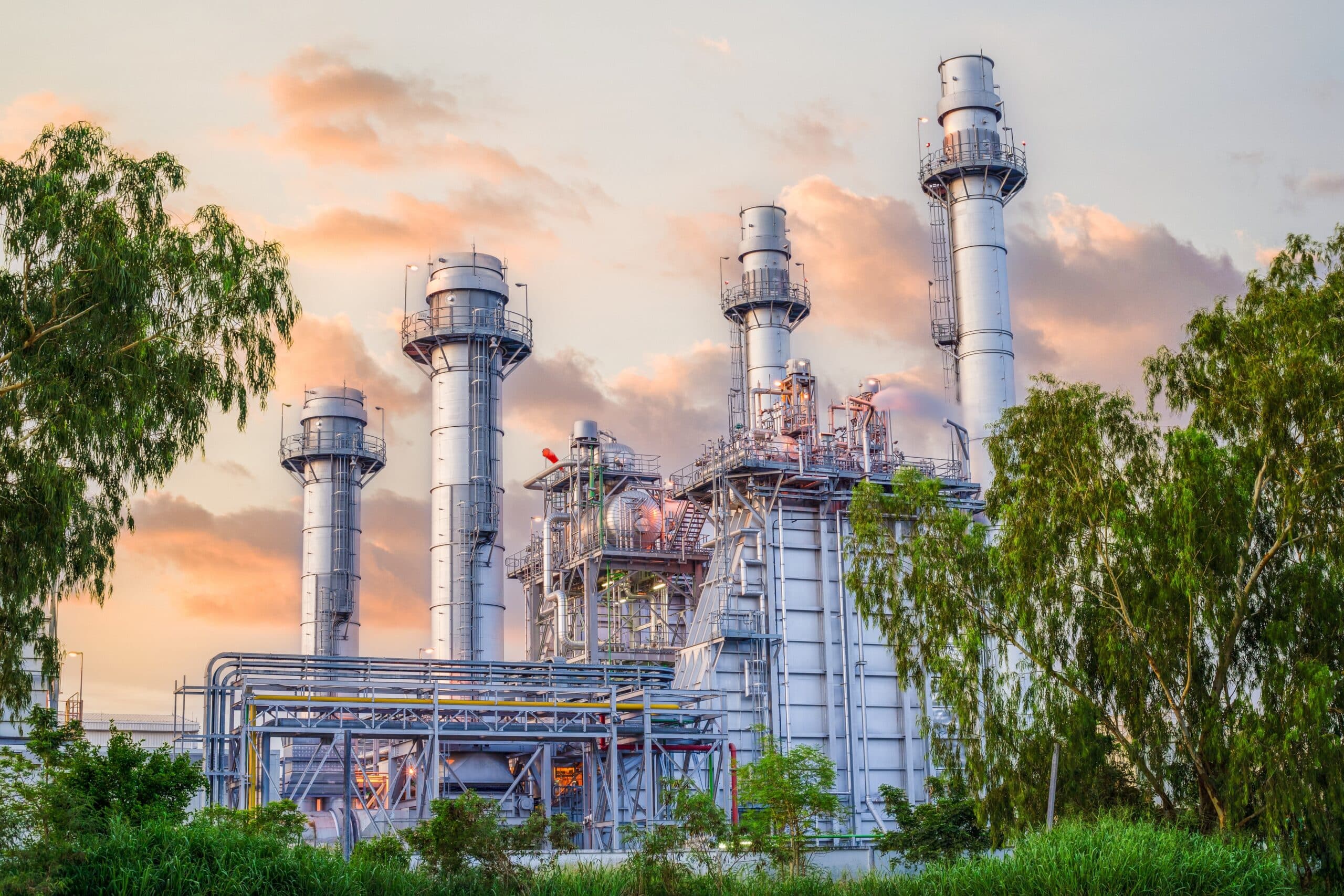A gas-fired power plant with four tall exhaust stacks at sunset, framed by trees.