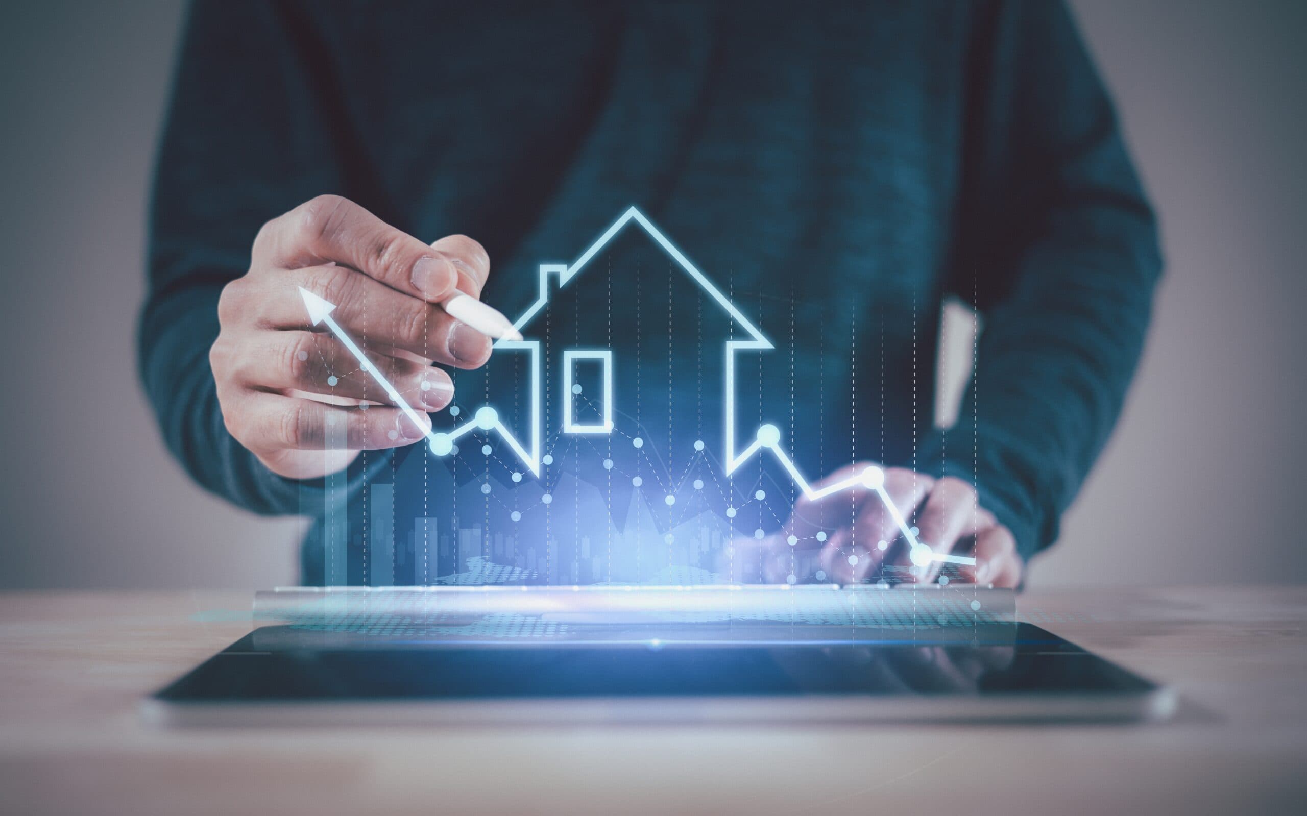 A person works at a tablet as a glowing outline of a house and rising and falling line charts project above the screen.